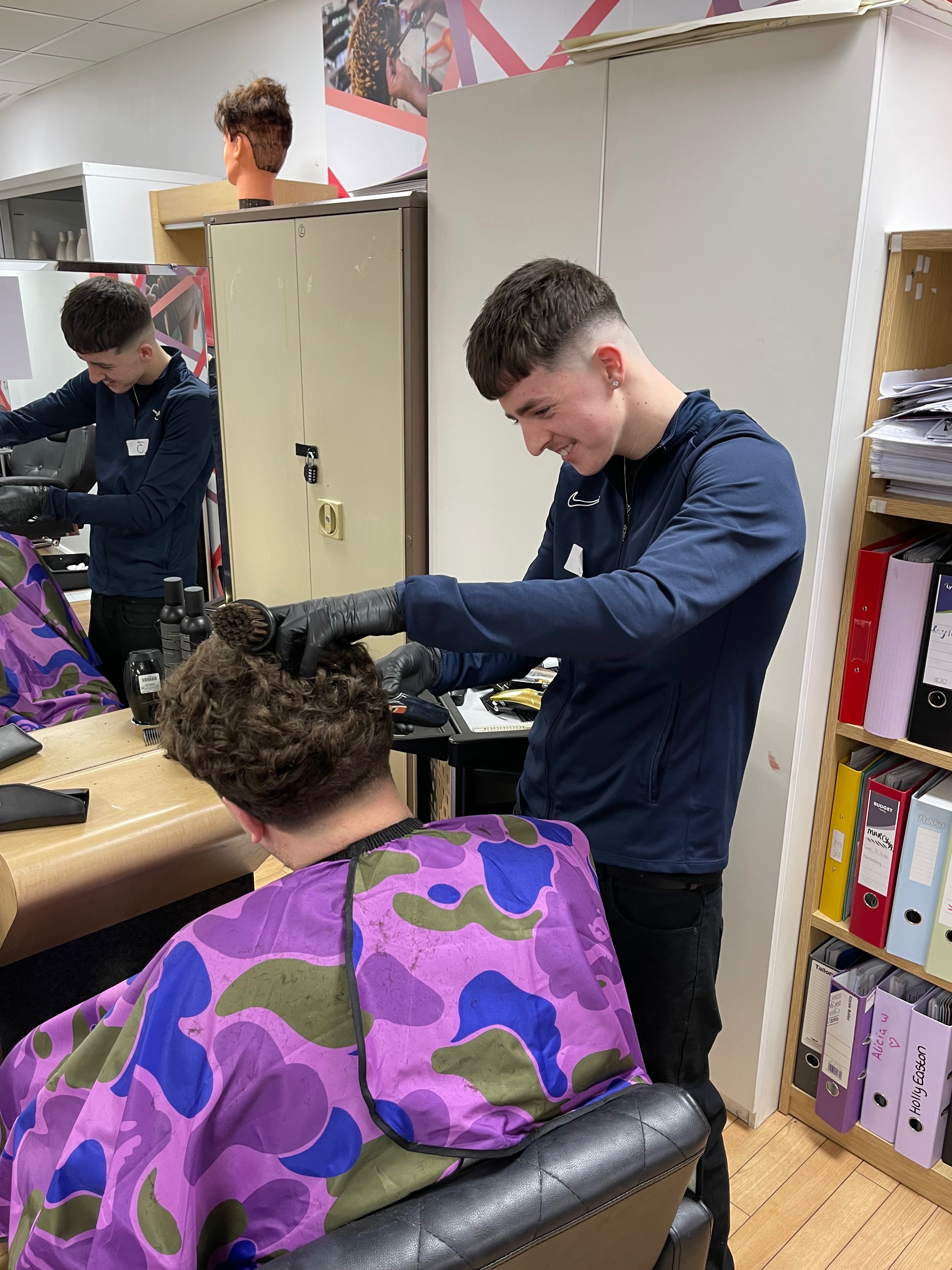 Corben stood to the side of a barbers chair, using a trimmer on the side of a clients hair